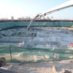 A long white crane looms over the Al Khayrien Tower worksite covered in building materials, dips in the surface, and construction workers.